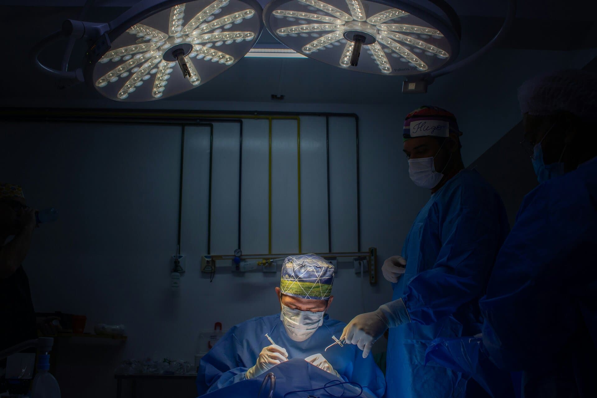 Surgical team in masks, gowns, and gloves completing a procedure as one surgeon hands medical equipment to another under two overhead lights.