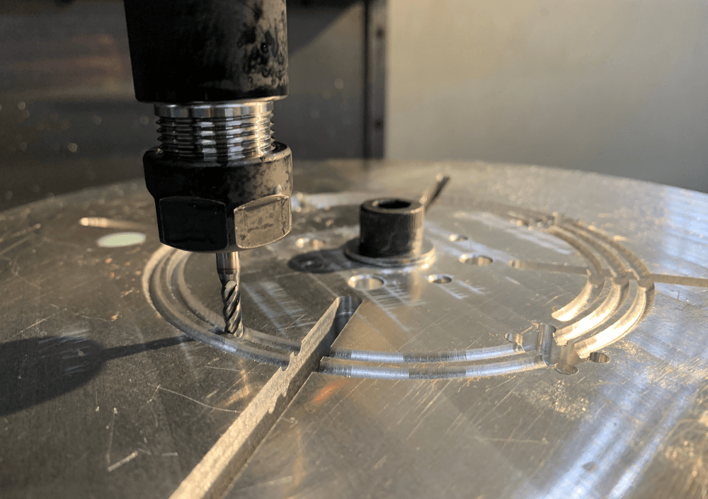 End mill cutting a curved slot into a round aluminum plate held on a machining fixture, with fine chips along the cut.
